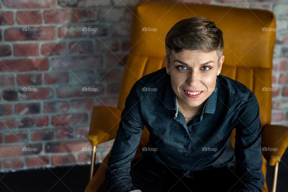 A business woman with a short haircut sits on a yellow armchair, a beautiful portrait with a look at the camera, beautiful eyes