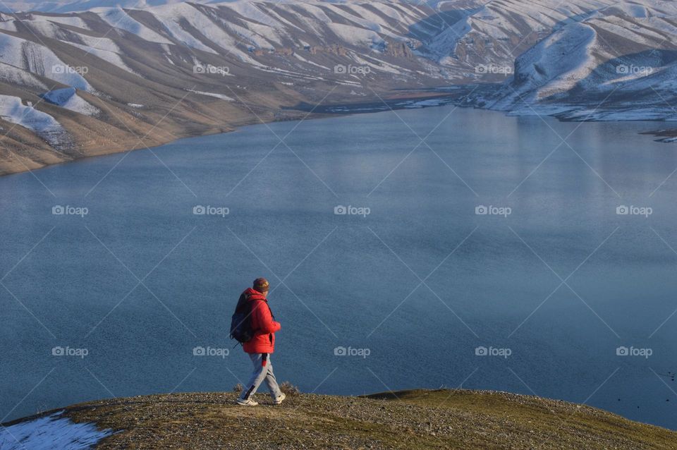 a man in a red jacket traveler walks along the shore of a mountain lake. in the background is a dark mountain lake and low peaks of mountains covered with snow