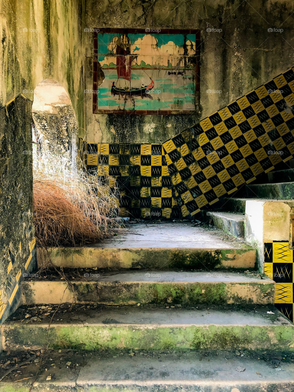 Plants grow through the broken window onto a neglected staircase, still decorated with moss stained tiles and murals, in an abandoned hotel - Portugal 