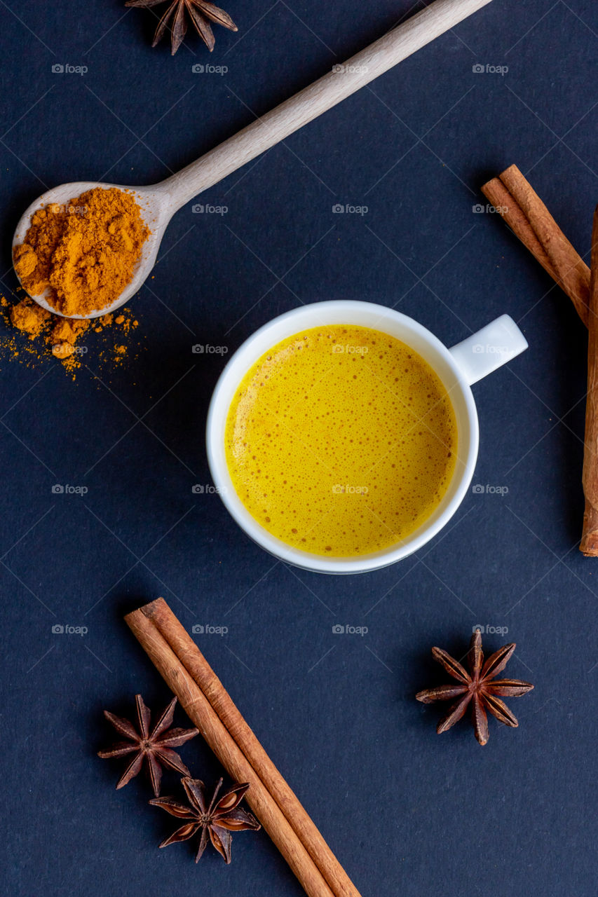 Golden milk in whie cup, cinnamon, anise and turmeric powder on dark background . Top view.