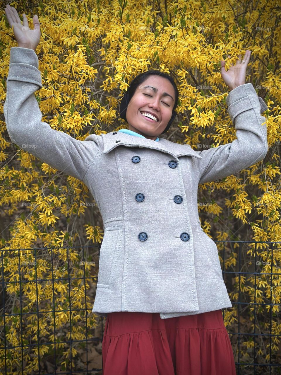 Smiling woman wearing coat, raising her arms against yellow flowers during spring.