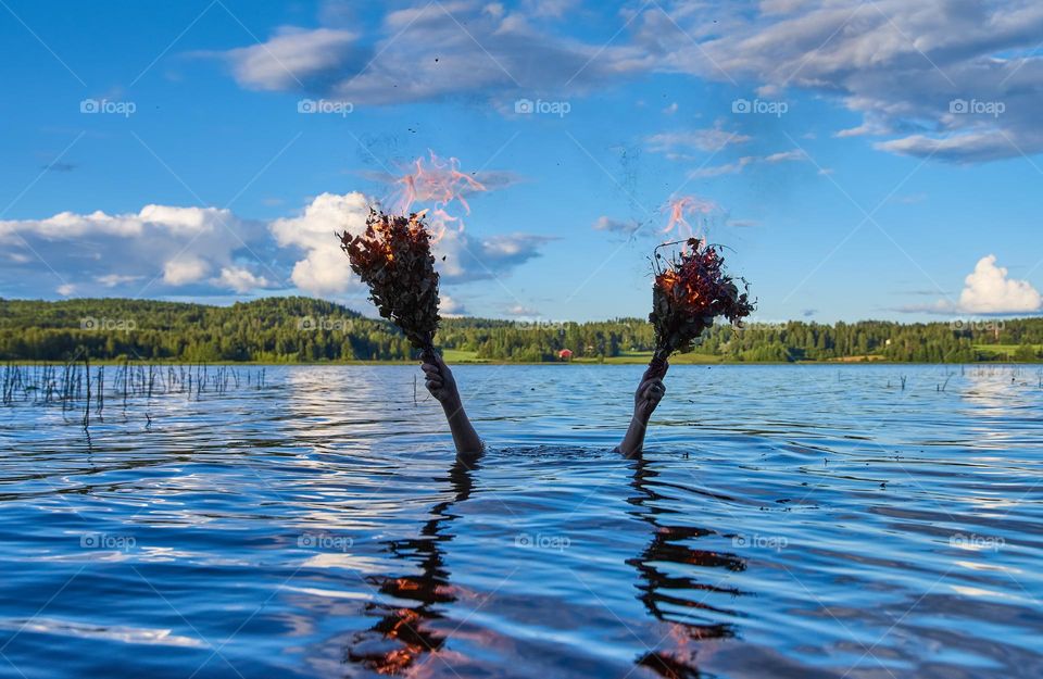 Submerged female holds two burning traditional Finnish bath whisks with her hands above water of a Finnish lake on a calm summer evening.