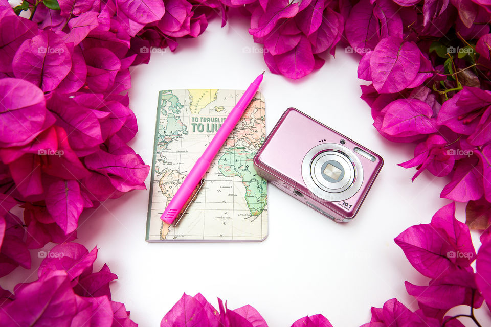 Even though we're stuck at home - we can plan and dream! I love exploring new places, taking photos and capturing memories. Image of camera, travel planning book, pink pen and flowers on a white background