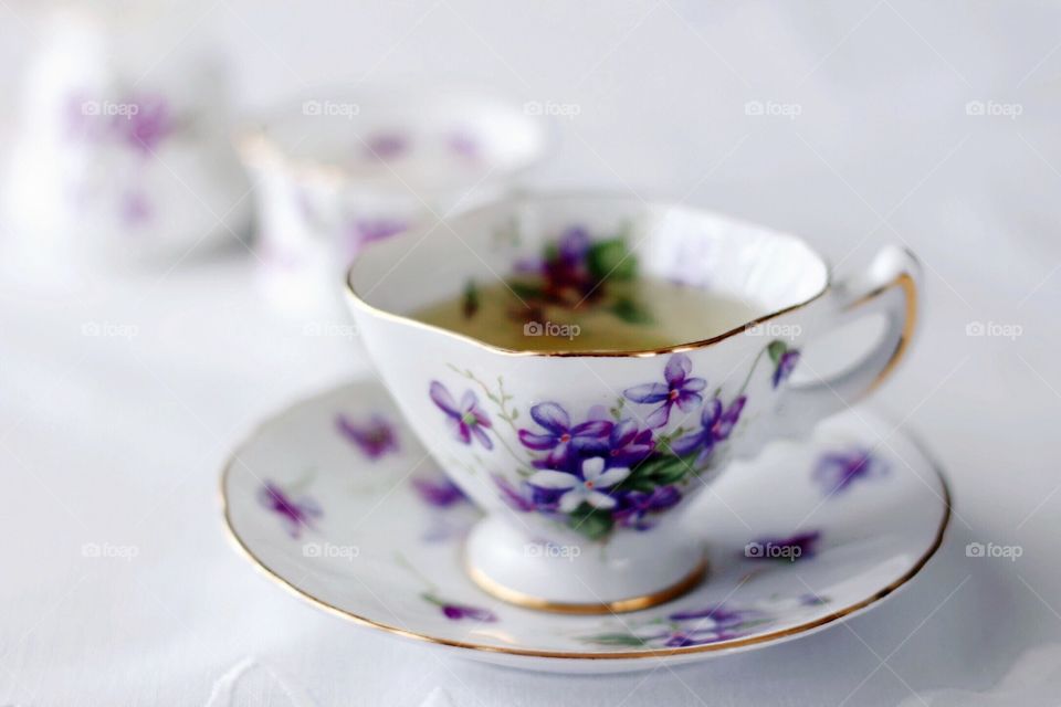 Green tea in a violet-patterned antique china teacup with saucer, sugar bowl and creamer on an antique white embroidered tablecloth 