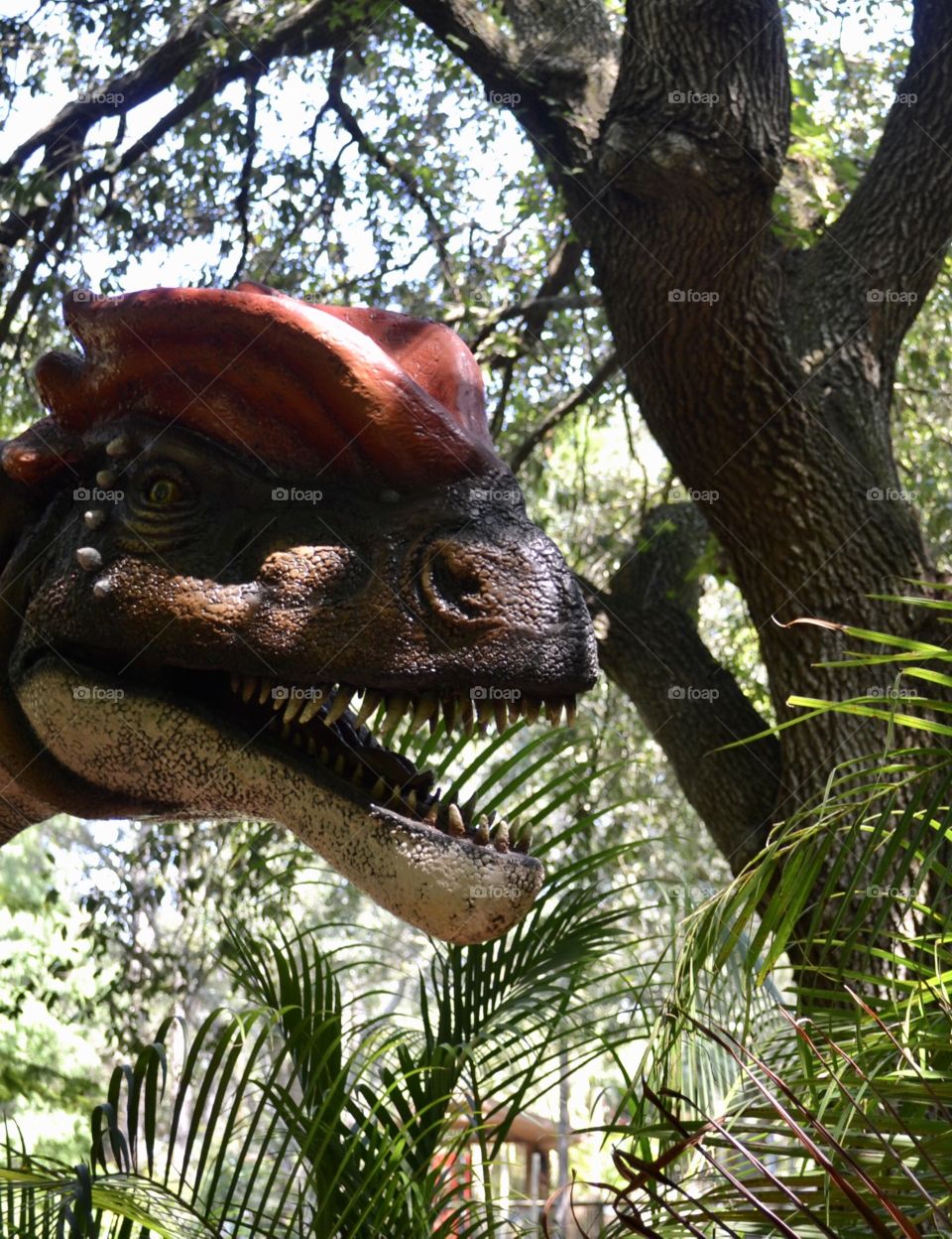 A closeup of a dinosaur head with its teeth showing. Trees in the background 