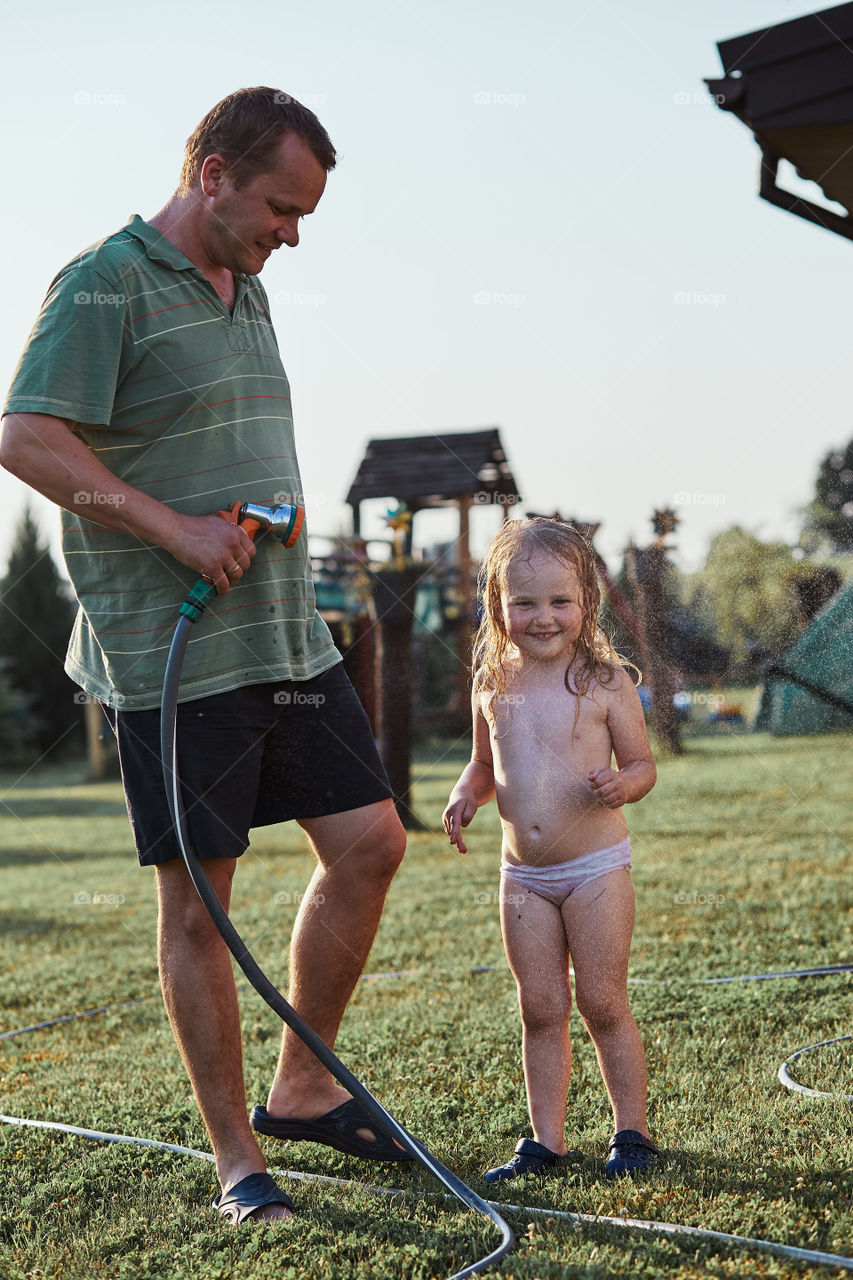 Little cute adorable girl enjoying a cool water sprayed by her father during hot summer day in backyard. Candid people, real moments, authentic situations