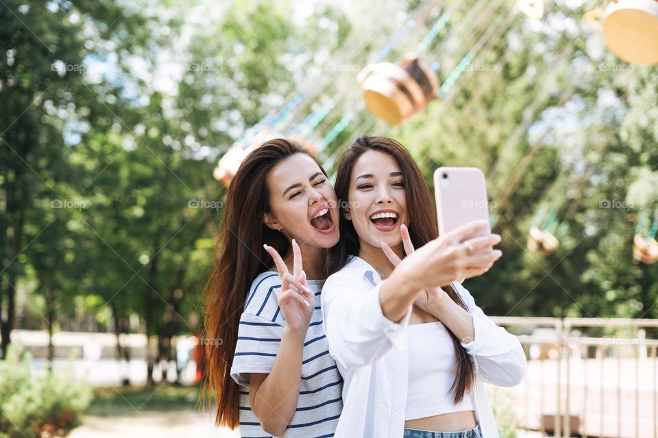 Young women with long hair friends taking selfie on mobile phone in their hands having fun at amusement park