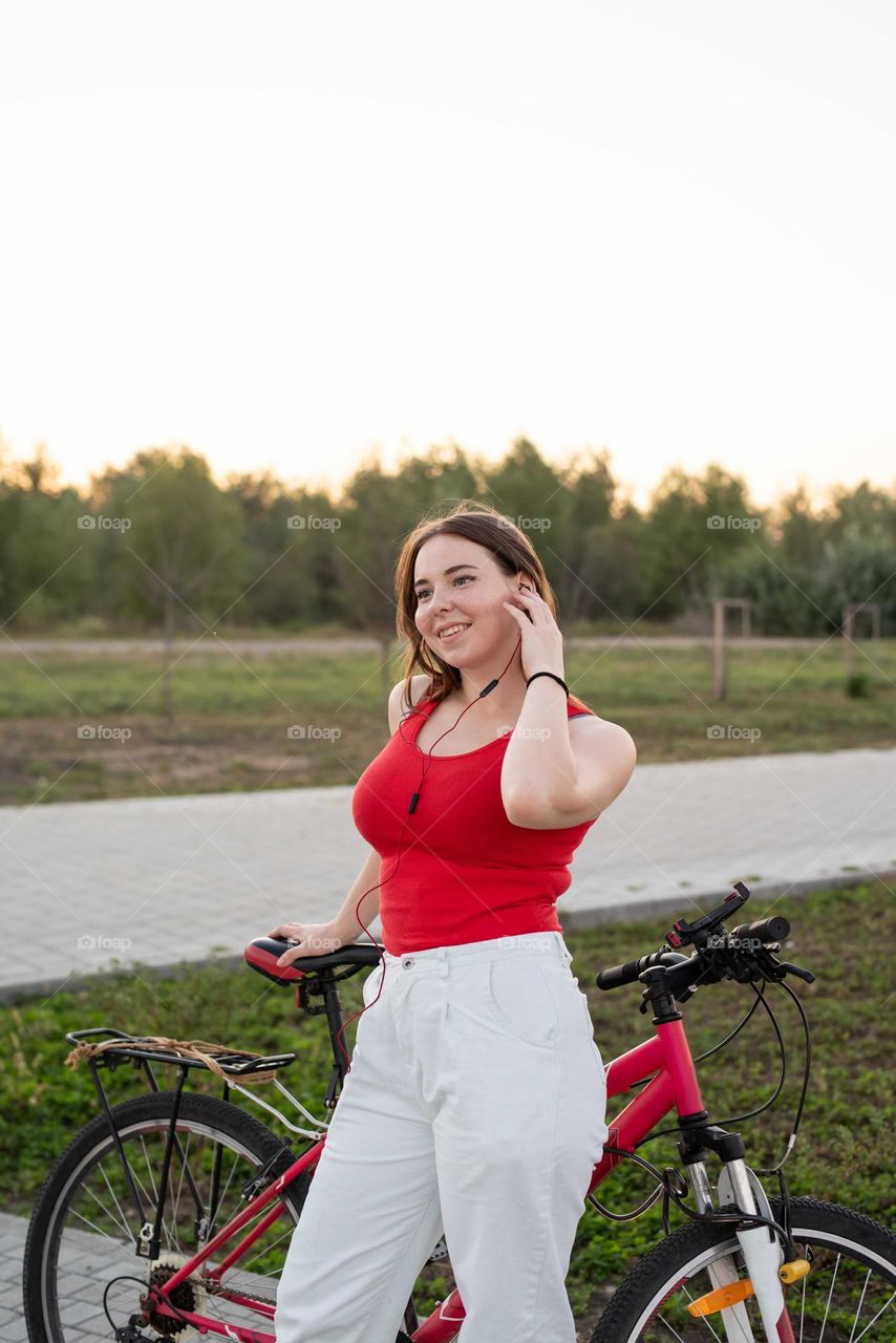 young smiling woman in red shirt and white pants listening to the music standing near her bicycle