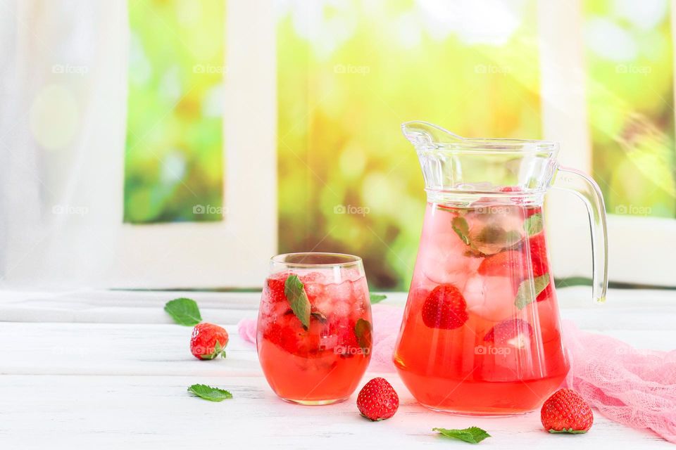 A cold strawberry drink with ice in a decanter and a glass with a gauze napkin lie on a wooden table against the background of a blurred window with a flowering field, close-up side view.
