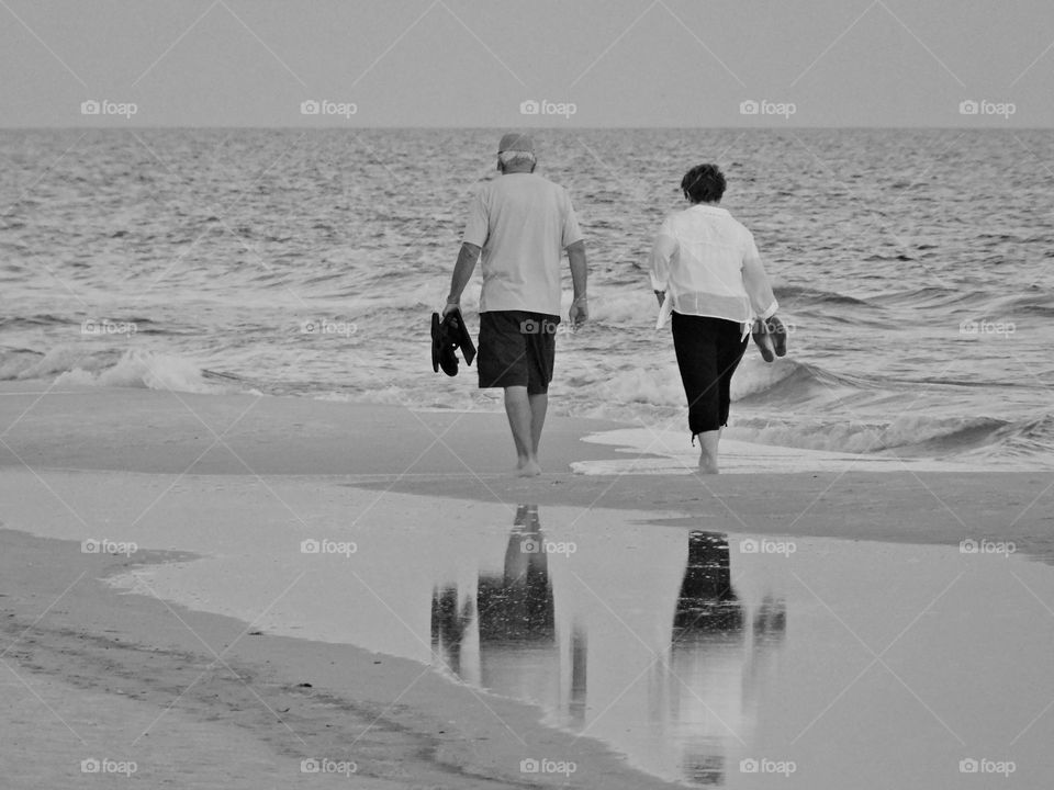 Beautiful B&W Seascape - A couple and with their shoes in their hand walk in the cool and soft white sand along the beautiful Gulf of Mexico. Their bodies reflect on shore. Florida is truly exquisite and one of the top attractions to the beach