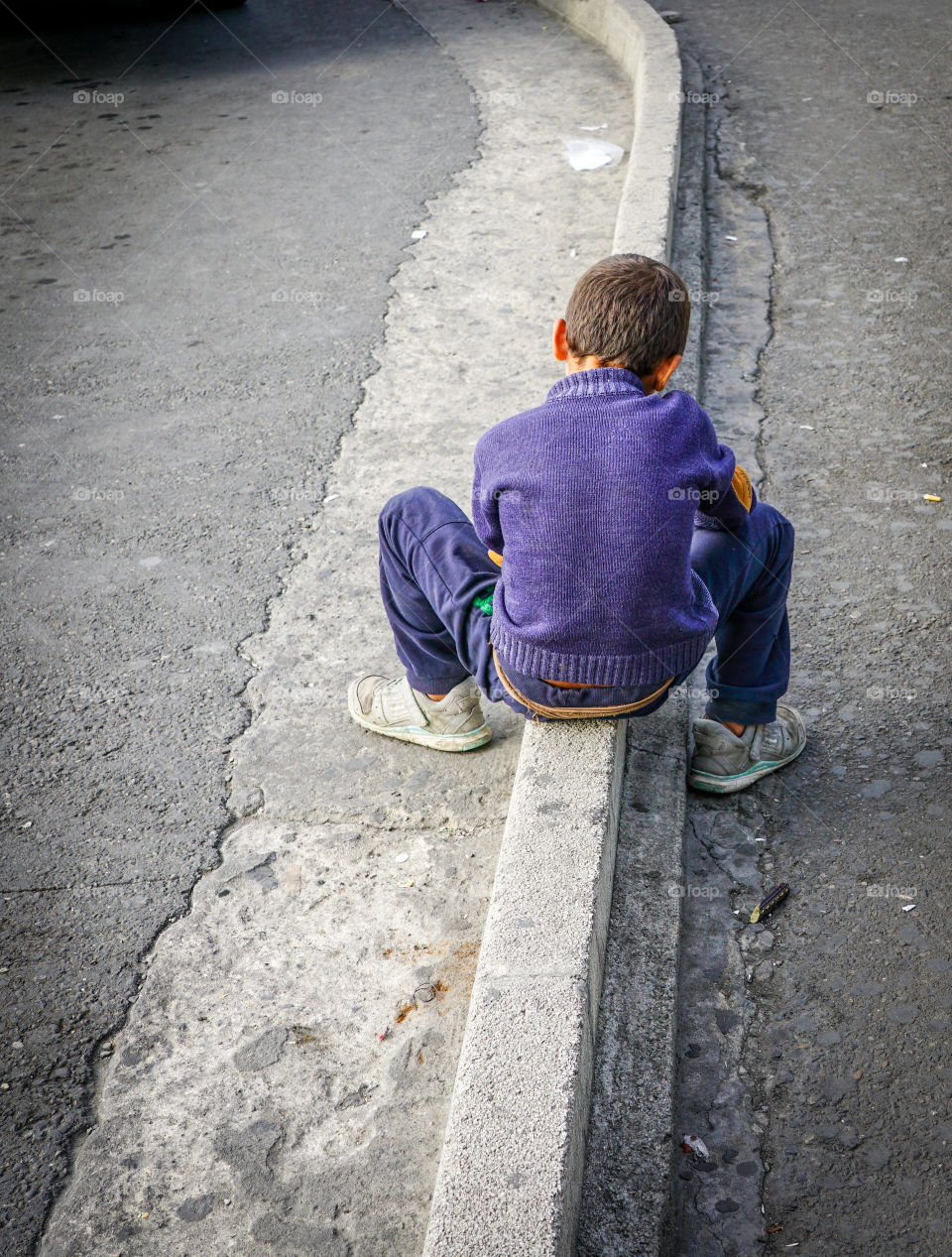 young boy in a purple sweater and blue jeans sits on the street border and think about future
