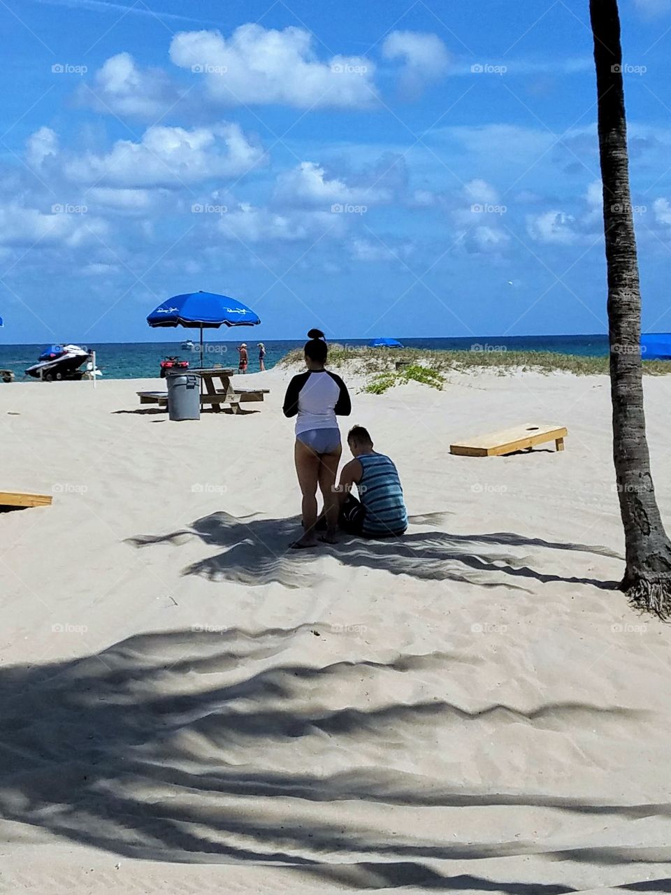Man & woman at the beach, so hot and sunny they're in the shade so they don't sunburn. The ocean is a far walk & there's no shade. Palm tree shadows are pretty on the beach. Sky is blue, beach is pretty.