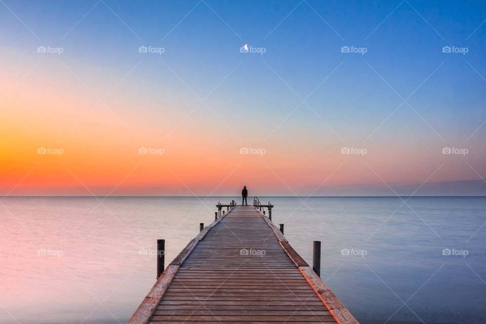 Jetty with silhouette standing in the end sunset sunrise time with moon in the sky. Blue and orange 