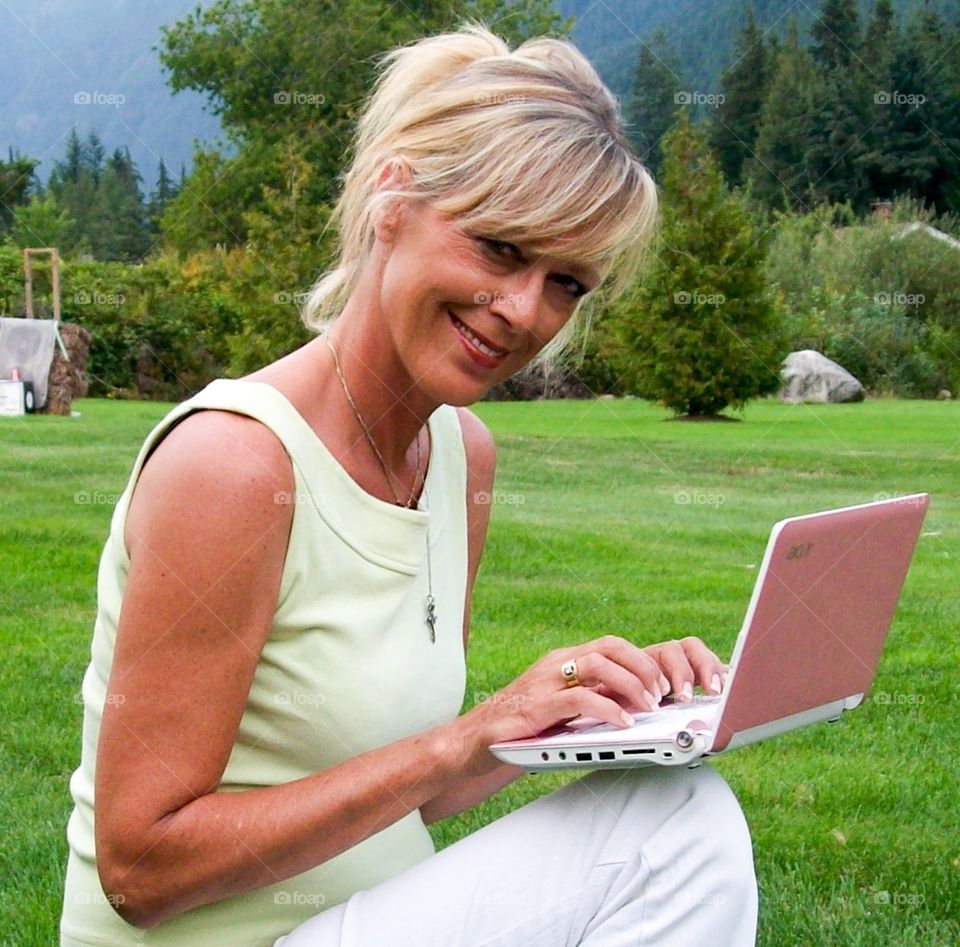 Blonde Woman with pink laptop squatting, kneeling down in grass 