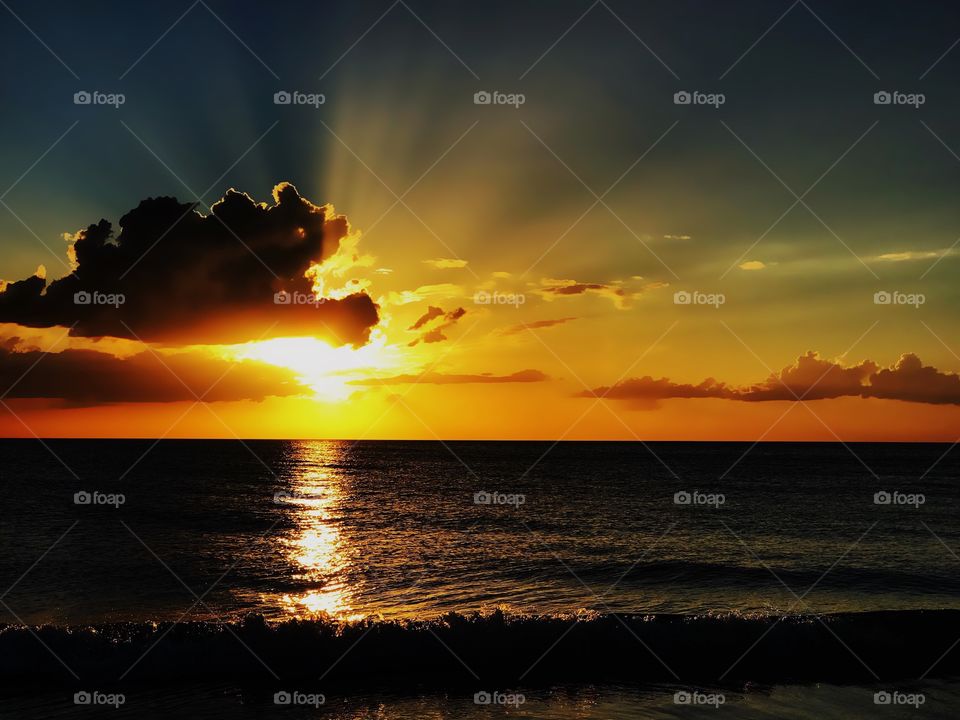 Nighttime sunset with a golden sun shining behind dark clouds over a dark ocean.