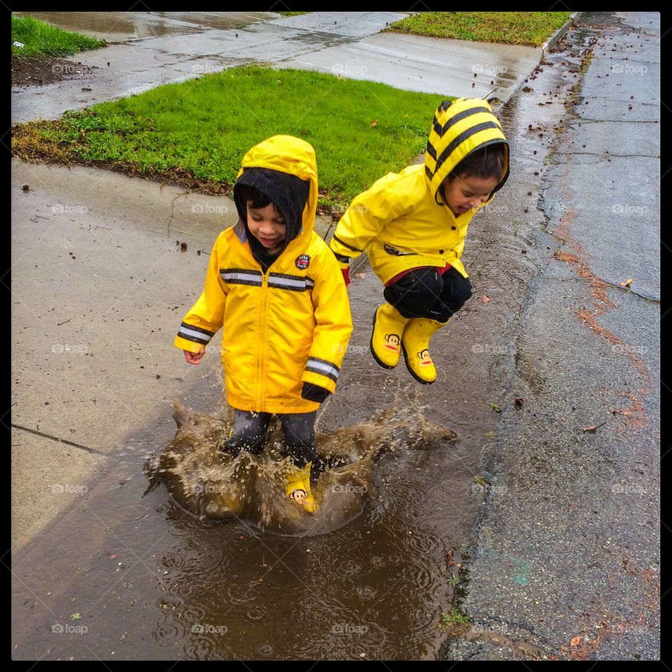 Jumping in puddles. Twin boys enjoy a rainy day, wearing yellow rain coats and boot while jumping in puddles. 