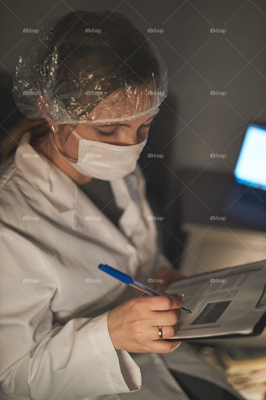 Doctor filling out a document. Hospital staff working at night duty. Woman wearing uniform, cap and face mask to prevent virus infection