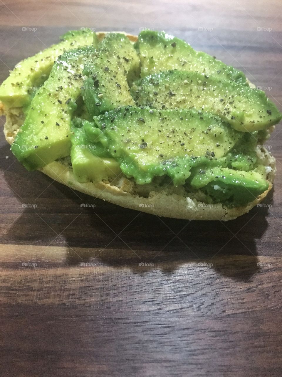 Avocado toast: sliced avocado on a toasted English muffin seasoned with salt and pepper atop a wooden plate.