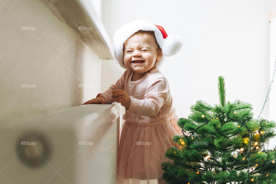 Cute baby girl in Santa hat in kitchen with Christmas tree at home 