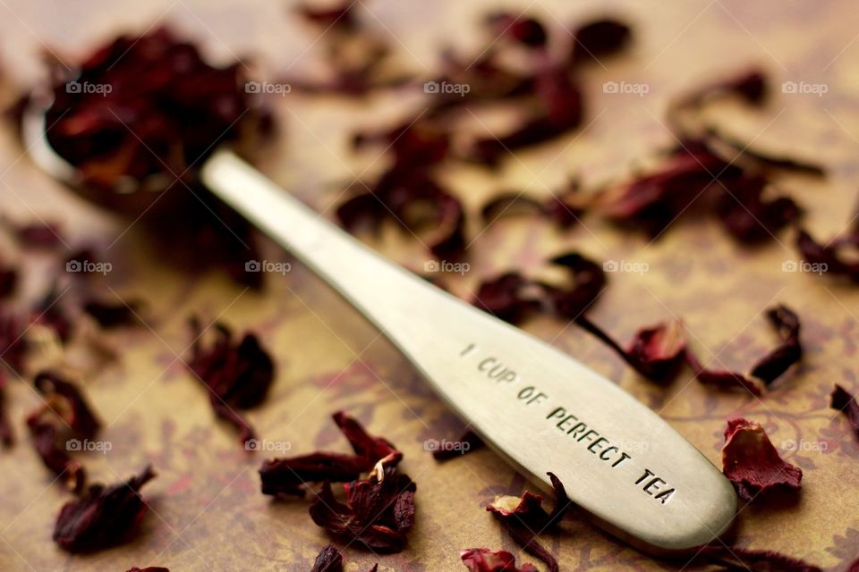 Angled view of a stainless steel measuring spoon imprinted with the phrase, “1 CUP OF PERFECT TEA,” filled with dried hibiscus petals on a brown and burgundy patterned surface with scattered hibiscus tea 