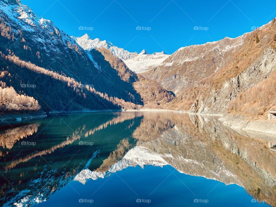 A breathtaking view of a crystal-clear lake framed by majestic mountains, with the sky reflecting on the water in a dazzling play of light and reflections. Nature here feels suspended in time, offering an atmosphere of peace and wonder.