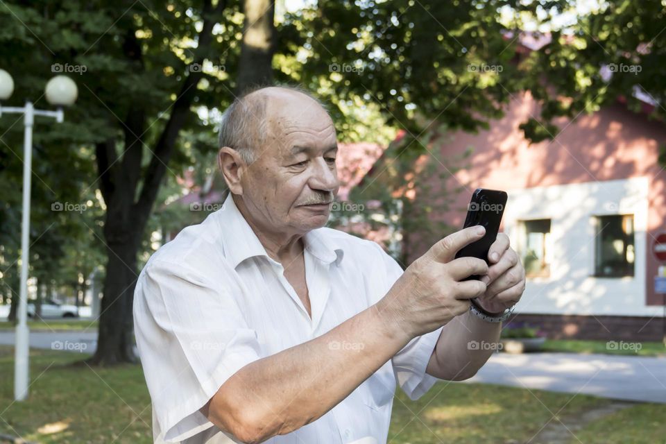 An elderly man walks alone in the park in the summer. A modern pensioner, businessman in a white shirt and trousers takes pictures with a camera in a mobile phone.