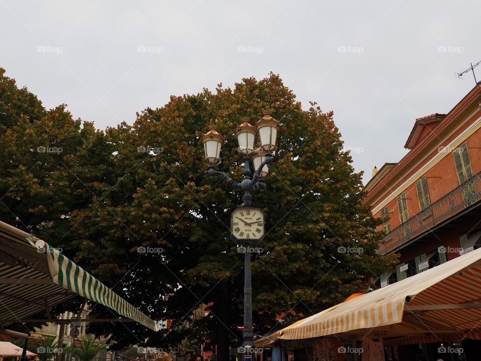 Ornate lamp post and clock surrounded by tree and colored building on the Cours Saleya in the old town of Nice, France.