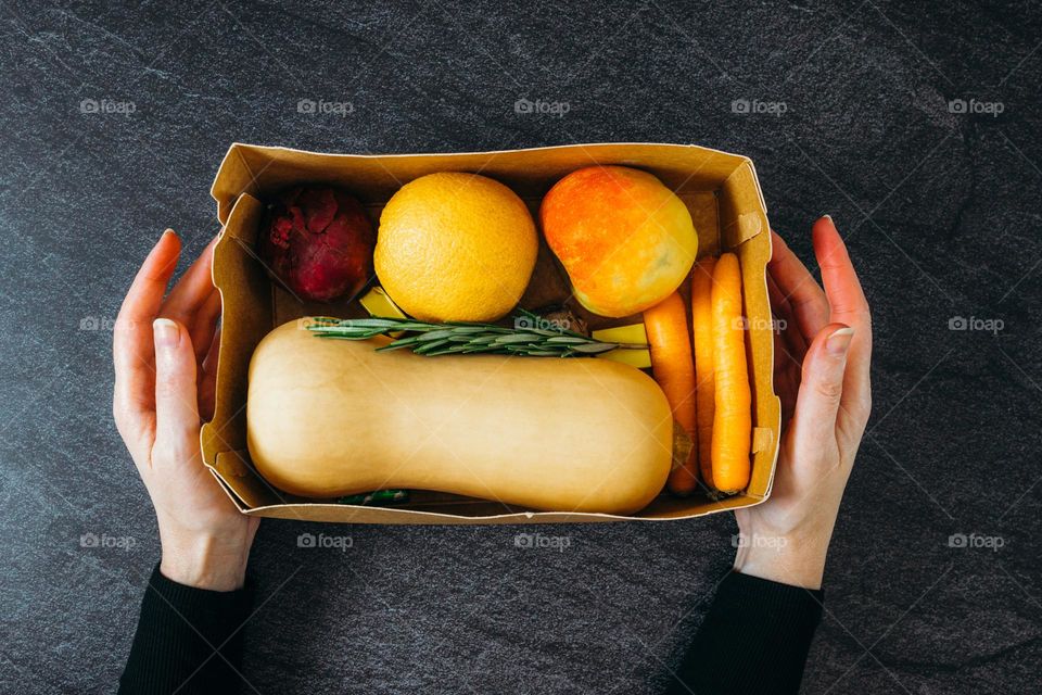 A young woman's hands hold a cardboard box of seasonal vegetables in a pumpkin soup set on a black stone table, Flt. Lay close-up.