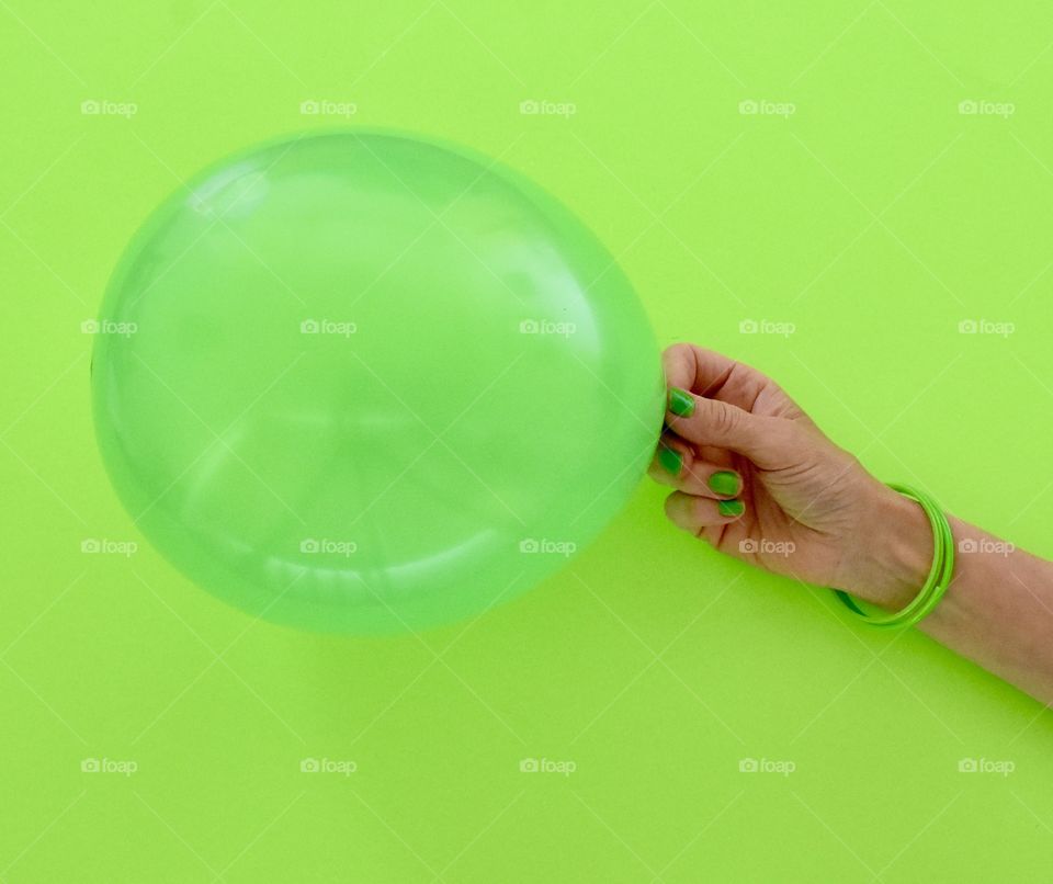 Green balloon against a green background held by a hand with a green bracelet and green nail polish 