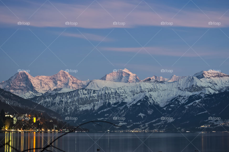 Mountain range (eiger, mönch & jungfrau) shortly after sunset with village lights reflecting in lake thun during winter.