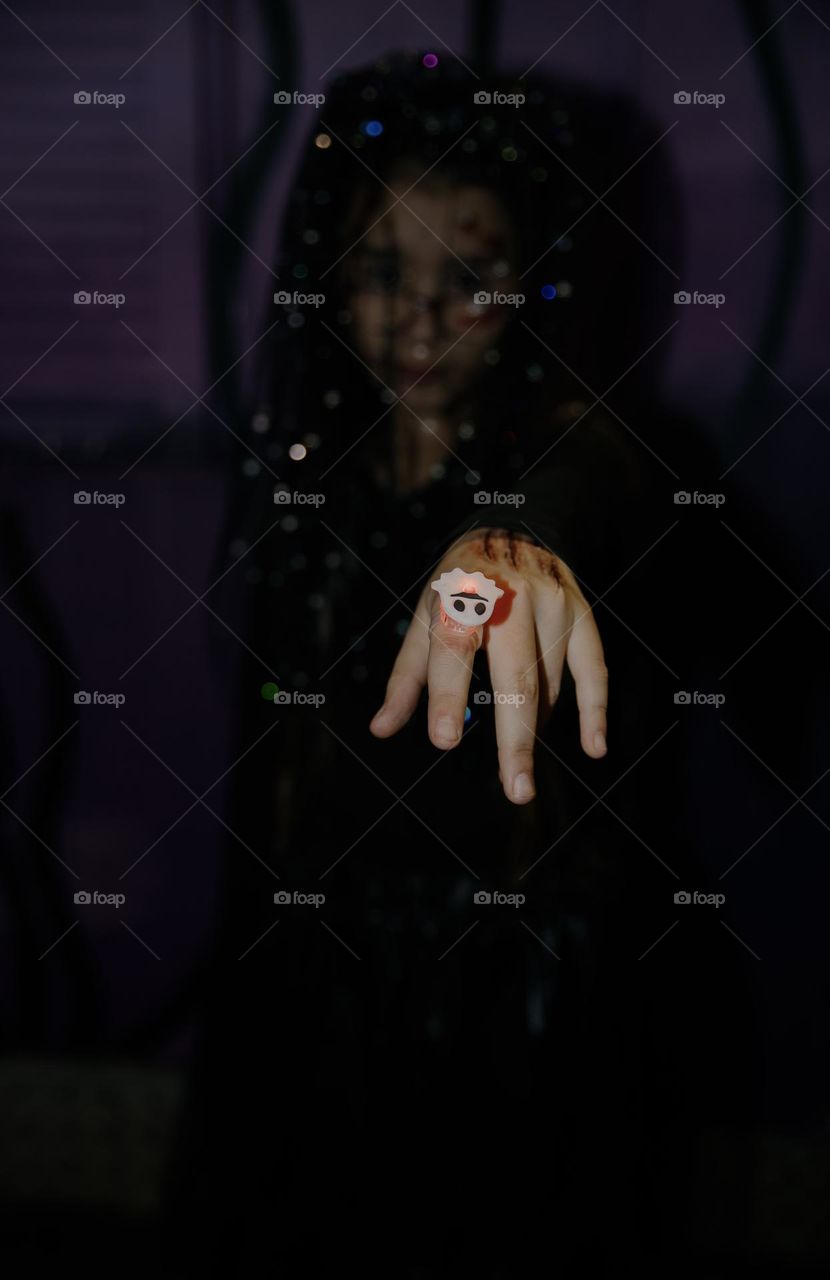 One little Caucasian girl in a dead bride costume extends her hand with a glowing ring in the shape of a white skull on a dark night on the street, close-up side view.