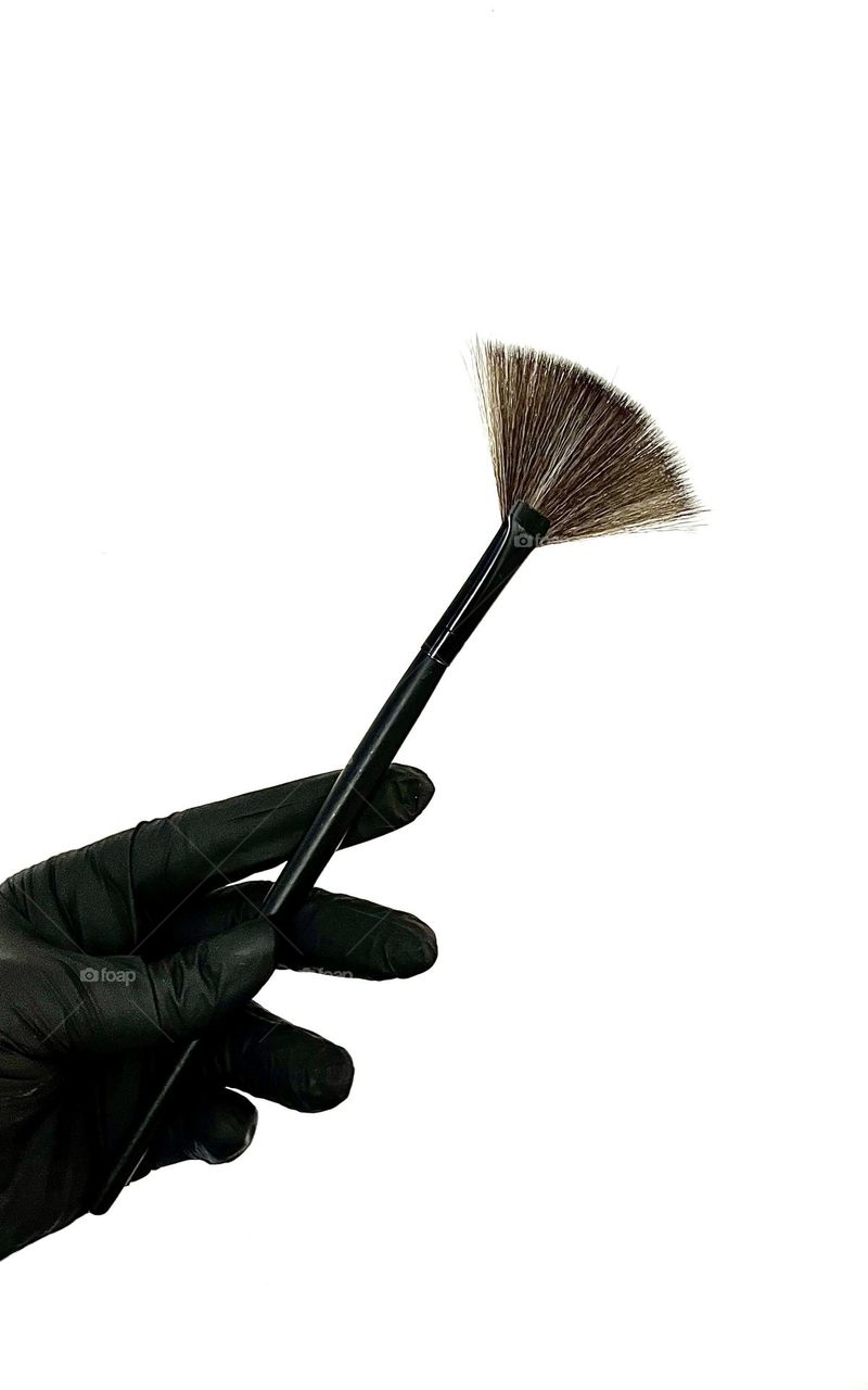 Hand in black nitrile glove holds brush for applying cosmetics on white background.