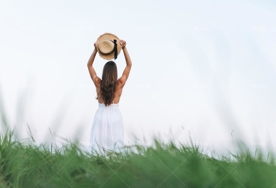Young beautiful carefree long hair woman in white dress with straw hat in sunset field, view from back. Sensitivity to nature concept