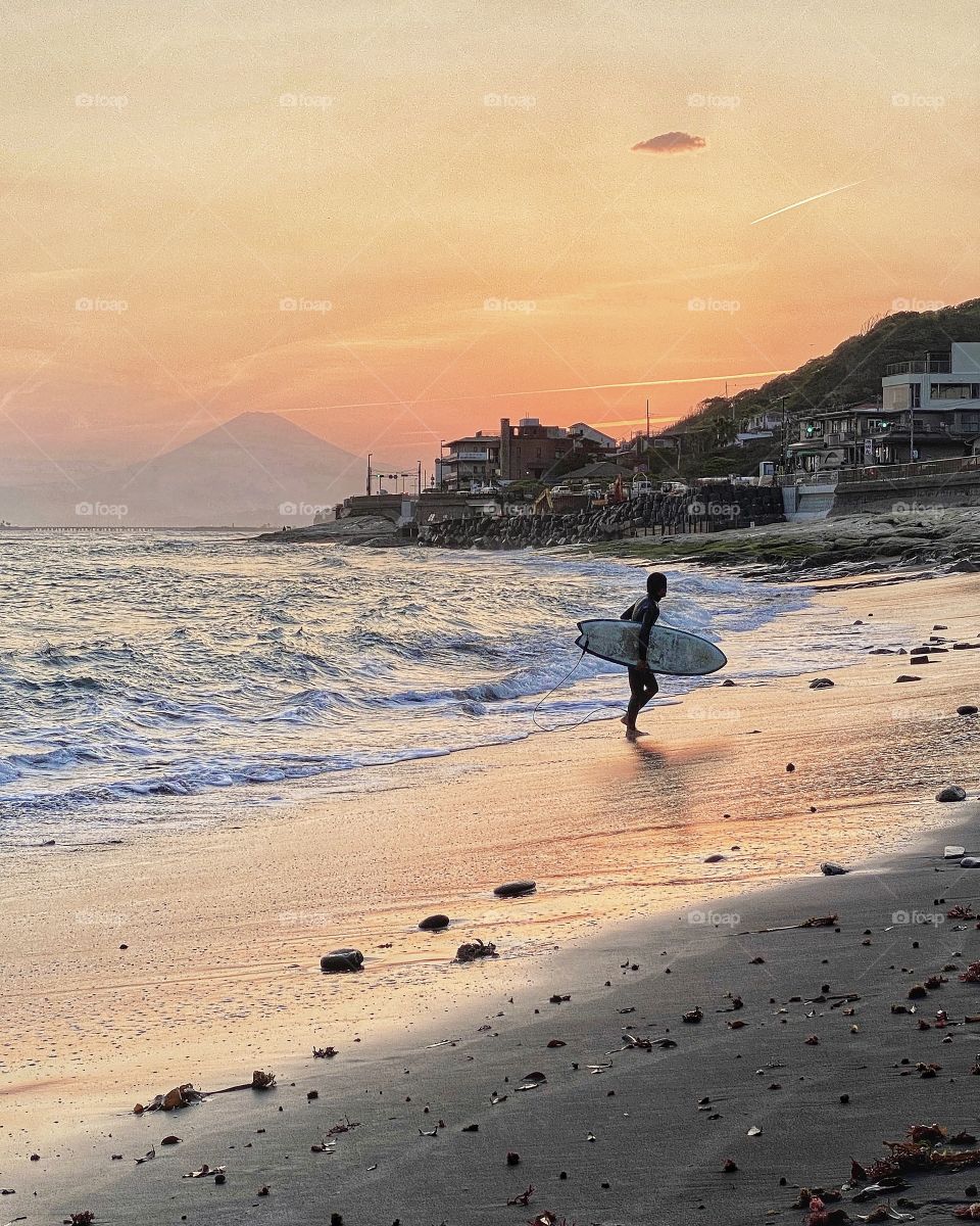 Surfer comes out of ocean with back drop of sunset colors and faded Mt Fuji in the distance.