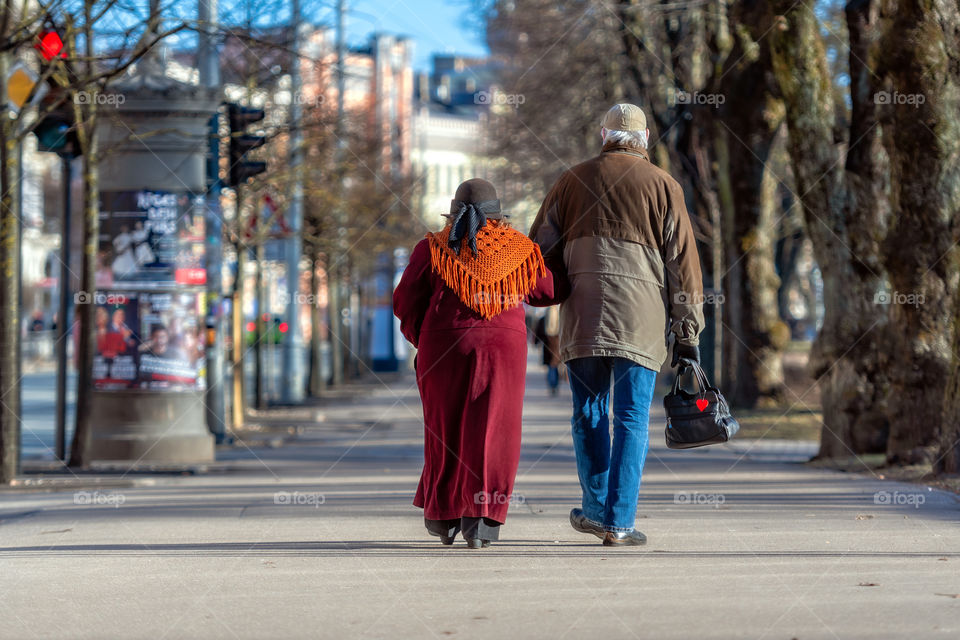 A couple of eldery people in hands are walking through the city street. Rear view.