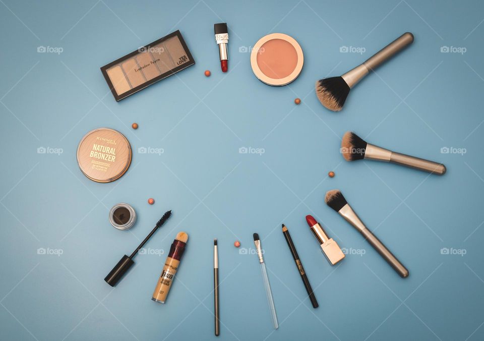 A cosmetics set of pallet of nude eye shadows, round face powder, red lipsticks, soft black eyebrow shadows, makeup brushes, mascara, eye lashes and foundation lie in a circular frame on a blue background with a copy of the space in the center, flat