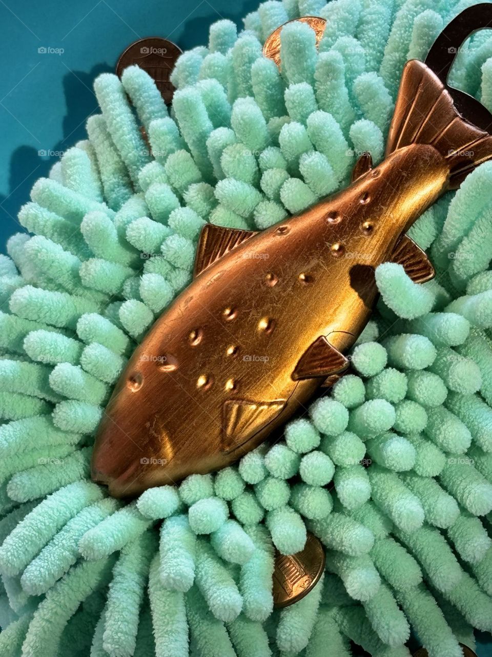 Copper coated fish shaped beer can opener lying in an aqua colored synthetic dust mop head with copper pennies on a teal plate 
