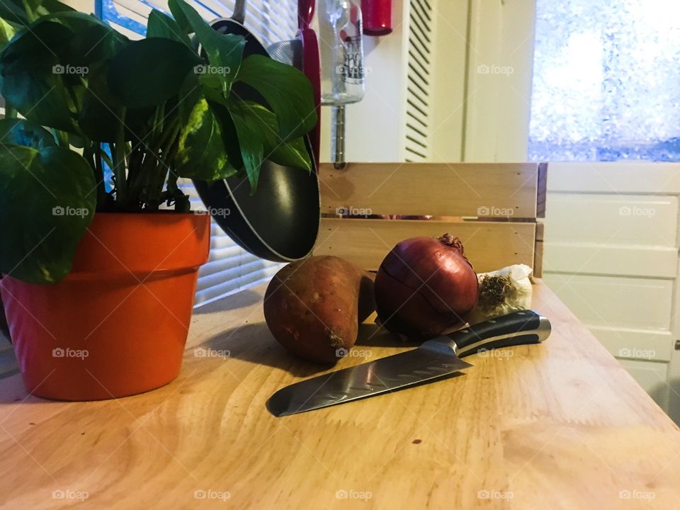 Fresh sweet potato, onion, and garlic on a cutting board next to a lush green indoor plan 