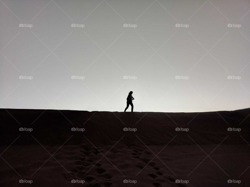 Woman (Girl) Walking Solo (Alone) Across the Sand Dunes of the Sahara Desert in Morocco at Dawn Just Before Sunrise (Backlit)