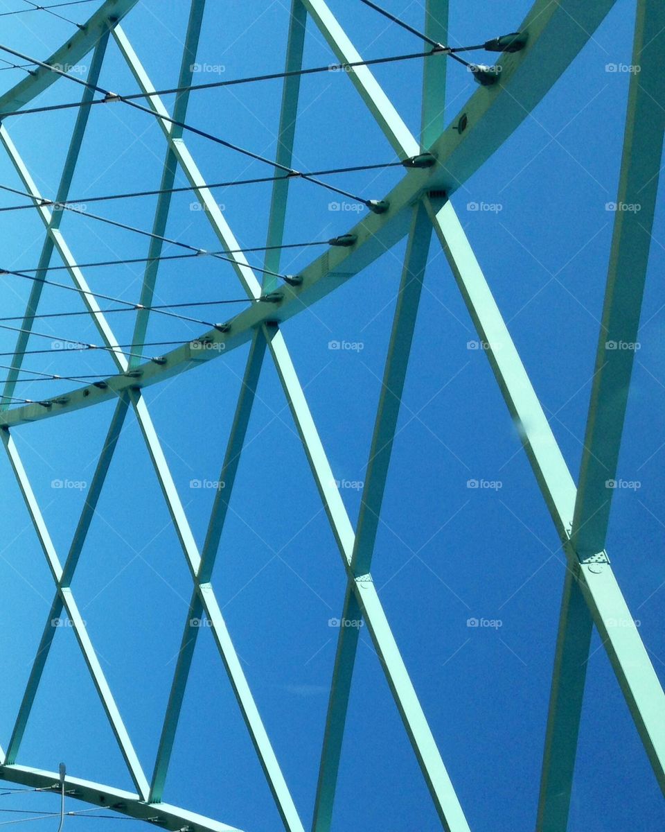 Bridge structure with steel bars painted green that cross one another in repetition, against a blue sky background. 