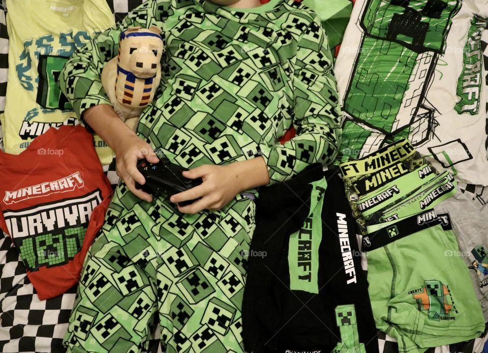 Kid with Minecraft pyjama holding Xbox controller and hugging Minecraft llama and many Minecraft clothes around him