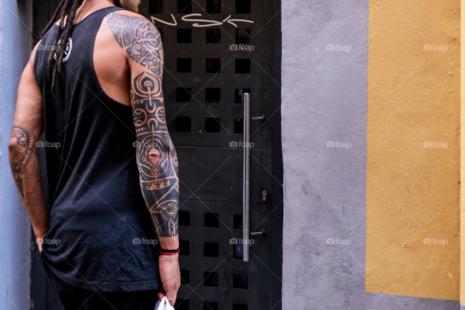 Esbelto hombre tatuado con figuras geometricas ante la puerta de restructures tambien geometrica de una viviendo pintada a rayas de colores.