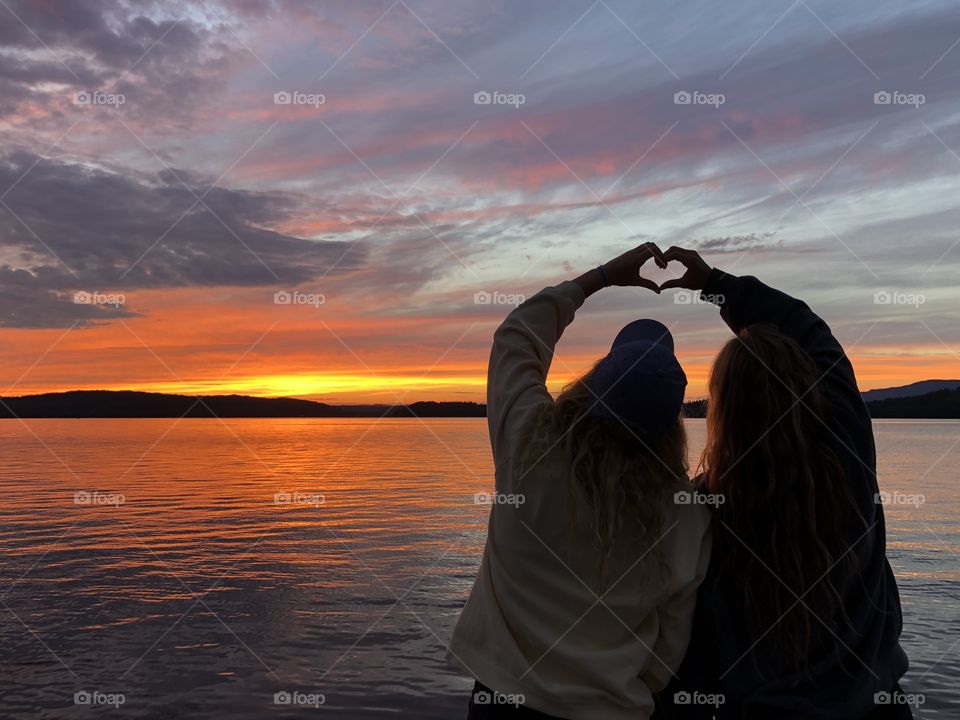 Two Girls Make a Heart Silhouette Against a Sunset Reflected on the Lake