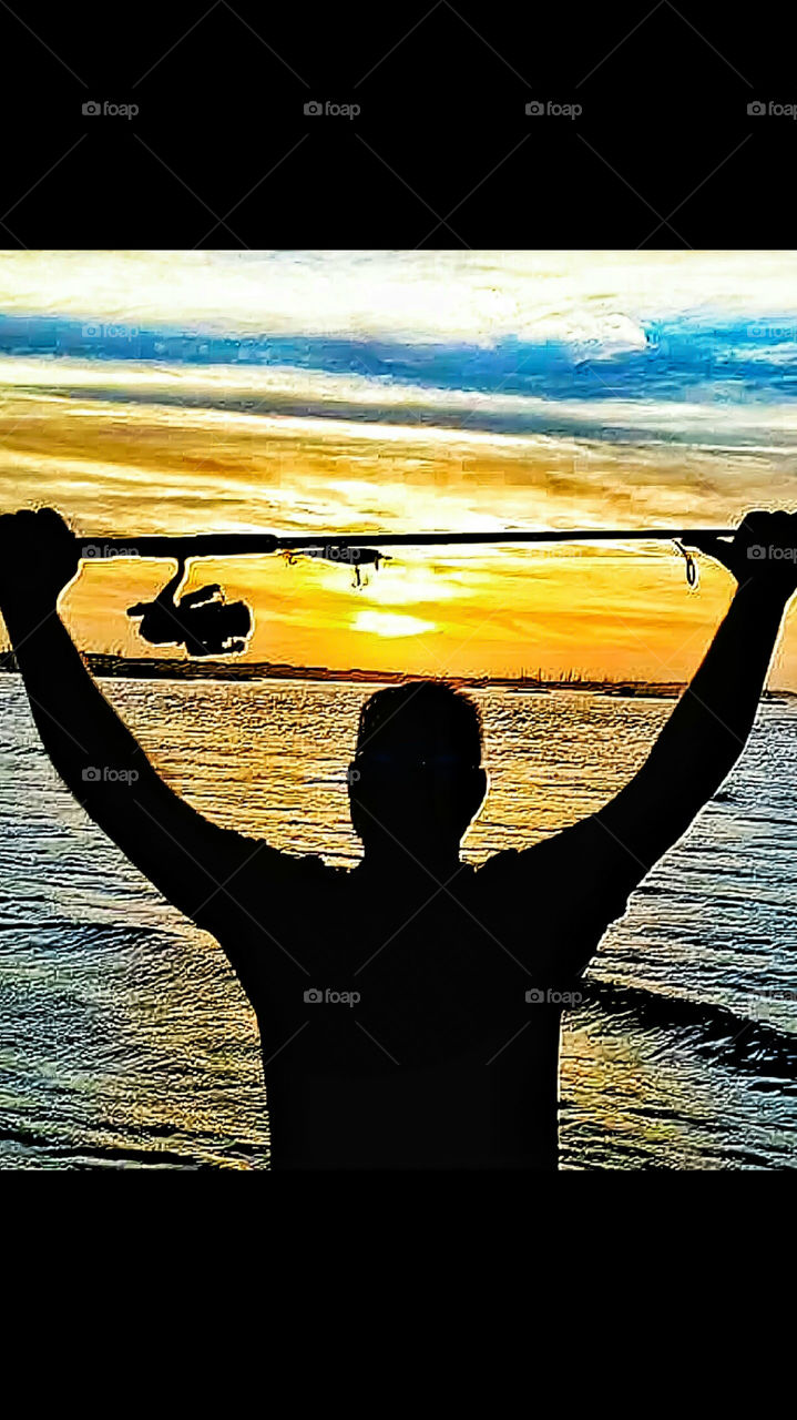 man holding rod and reel overhead with water and sun rise in background