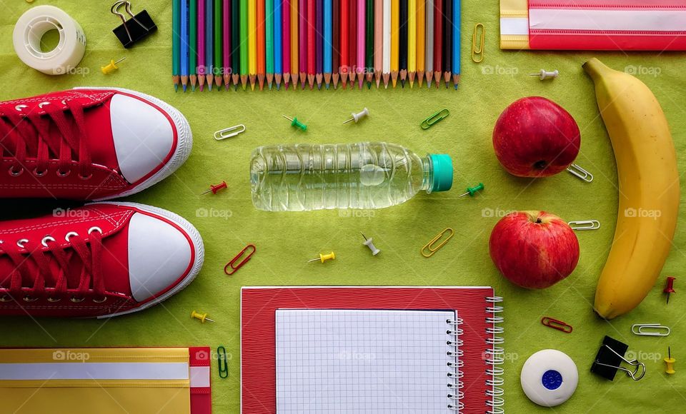 Concept School! School accessories with healthy snack and the bottle of water. Red and yellow folders in the corners, sneakers, a banana, two apples, a bottle of water, buttons, colored pencils, paper clips, scotch tape, eraser, spiral copybooks✏️🏫