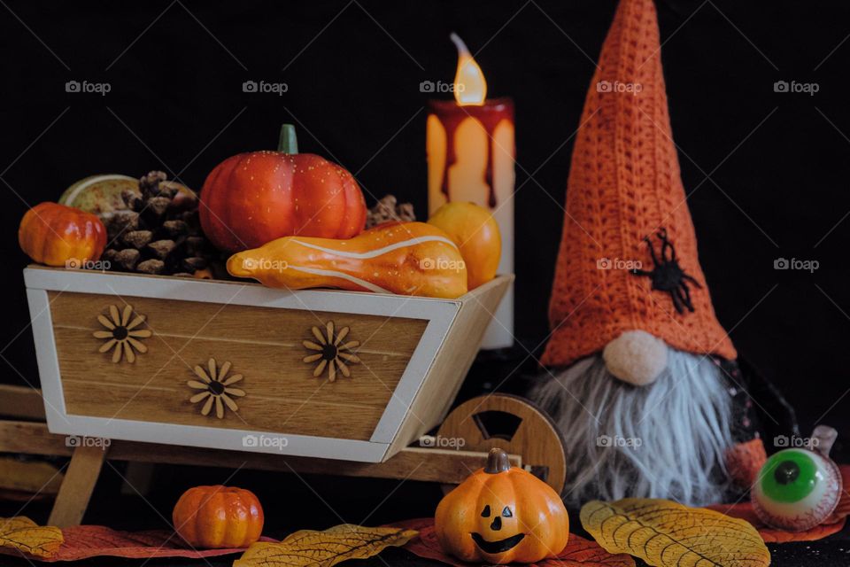One wooden homemade wheelbarrow full of decorative pumpkins and pinecones with Halloween gnome toy, plastic candle with colorful foliage stand on a table against a black wall, close-up side view.