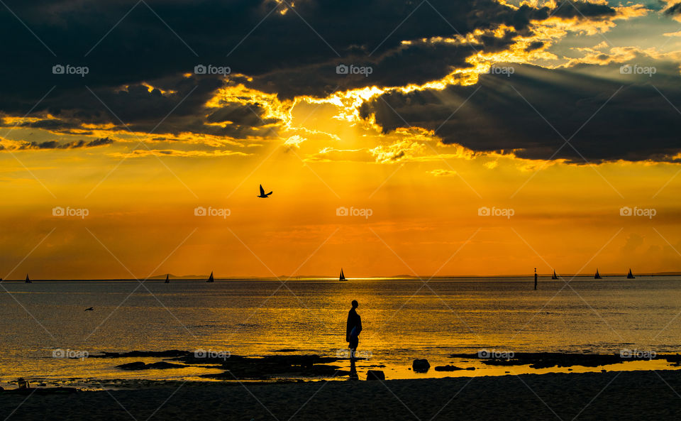 silhouette of man standing on beach as the sun begins to set, casting golden rays and color across the sky