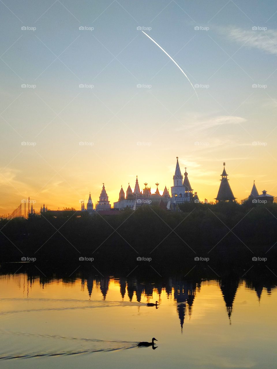The river is orange from sunset on which ducks swim.  The domes of the castle are visible across the river among the green trees.  The sun is setting behind one of the domes.  The white stripe of the aircraft taking off is visible in the blue sky