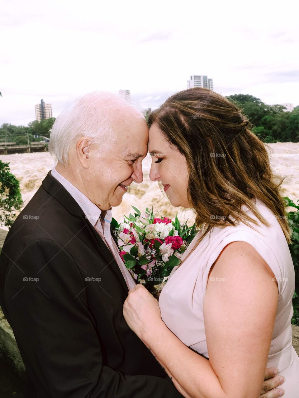 A groom and a bride with a flower bouquet, at the Piracicaba River, holding each other tight.