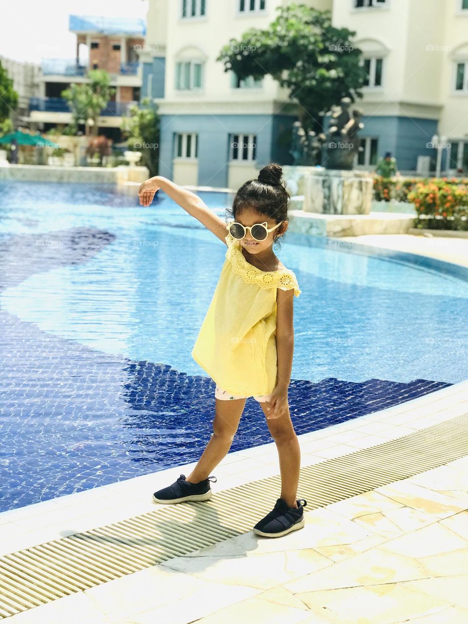 A girl in a yellow summer outfits and cooling glasses stands near swimming pool with a pose.